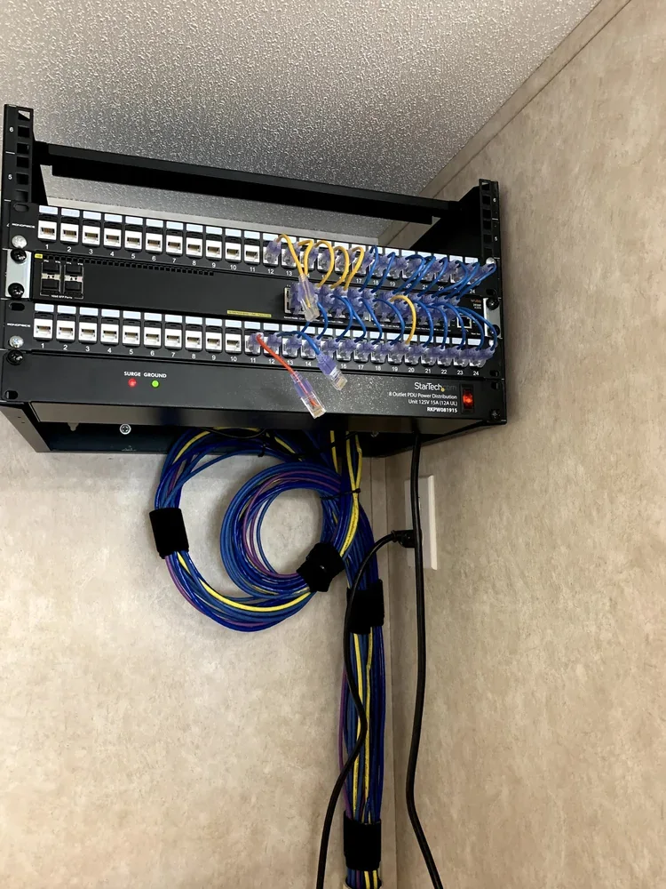 Small wall rack with numbered patch panels, a switch and power distribution above bundled blue and yellow cable